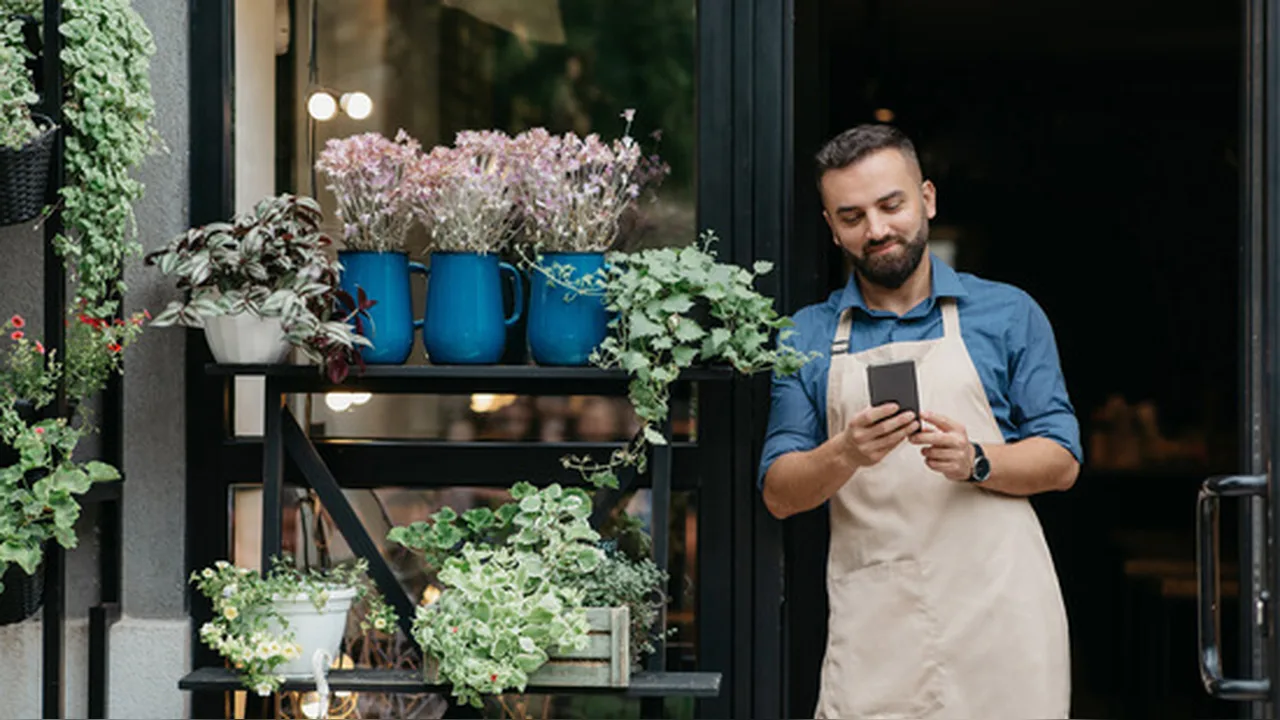 A small shop owner checking a phone beside products on a shelf